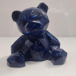 May include: A dark blue geometric teddy bear figurine with lighter blue flecks. The bear is in a seated position, with its arms and legs extended. The form is made up of angular planes. The figurine is on a white surface.