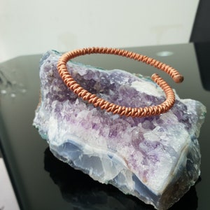 May include: A copper wire bracelet with a twisted design. The bracelet is resting on a purple and white amethyst crystal.