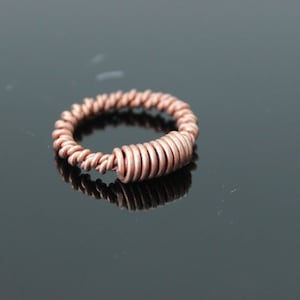 May include: A copper-colored ring with a spiral design. The ring features a tightly wound wire pattern around the band, with a thicker, coiled section on one side. The ring is displayed on a reflective black surface.