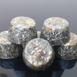 Peut inclure: Cinq savons ronds, gris, noirs et dorés avec des paillettes dorées et blanches. Les savons sont empilés les uns sur les autres.