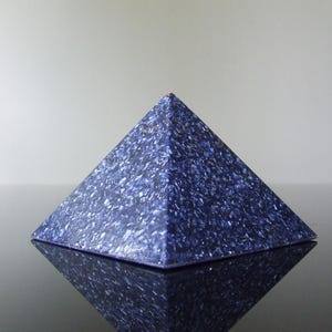 May include: A blue pyramid with a textured surface. The pyramid is made of a dark blue material with lighter blue and white flecks. The pyramid is sitting on a reflective surface.