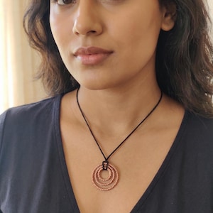 Könnte beinhalten: Kupferfarbene Anhängerkette mit drei ineinander verschachtelten, strukturierten Ringen. Die Ringe hängen an einer schwarzen Kordel. Die Halskette wird vor einem dunklen Hintergrund getragen.