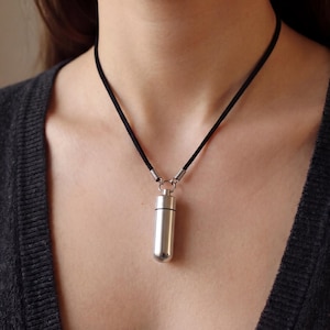 May include: A silver cylindrical pendant necklace with a black cord. The pendant is a small, capsule-shaped container. The necklace is worn against a dark grey cardigan.