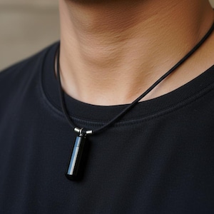 Op de afbeelding: Zwarte cilindrische hangerketting. De hanger hangt aan een zwart koord en is gemaakt van een glanzend, donker materiaal. De ketting wordt gedragen over een zwart T-shirt.