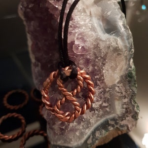 Puede incluir: Un collar con un colgante de cobre con un diseño de cuerda trenzada, colgando de un cordón negro. El colgante se muestra en una geoda de amatista púrpura y blanca.