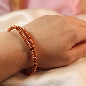 Pode incluir: Uma pulseira cor de cobre com um design em espiral. A pulseira é feita de arame enrolado e é usada no pulso. A pulseira tem uma cor marrom-avermelhada quente e parece ser feita à mão. O fundo é de cor neutra.