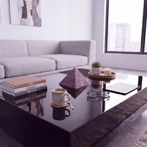 以下が含まれることがあります： モダンなリビングルーム。低い黒いコーヒーテーブルには、本、コーヒーカップ、装飾的なピラミッド、植物とキャンドルが入った小さなトレイが置かれています。背景には明るい色のソファがあります。