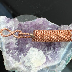 May include: A copper wire wrapped pendant with a twisted wire loop. The pendant is resting on a purple amethyst geode.