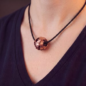 Könnte beinhalten: Kupferfarbene Halskette mit einem kugelförmigen Anhänger mit einem strukturierten, blattartigen Design. Der Anhänger hängt an einer schwarzen Kordel. Die Halskette wird über einem dunkelblauen Shirt getragen.