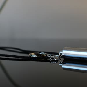Puede incluir: Un colgante cilíndrico plateado con extremos redondeados, sujeto a un cordón negro y un pequeño llavero. El colgante está hecho de metal brillante y refleja la luz. Probablemente sea un collar.