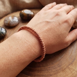 Puede incluir: Un brazalete en espiral de color cobre. El brazalete se lleva en la muñeca, con la mano apoyada sobre una superficie de madera. Varias piedras pulidas son visibles en el fondo, añadiendo un elemento natural a la composición.