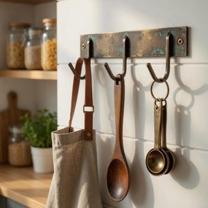 May include: Aged brass kitchen hook rack with three hooks, holding a tan apron with brown leather straps, a wooden spoon, and a set of brass measuring spoons. The rack is mounted on a white wall.