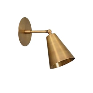 以下が含まれることがあります： 真鍮色の壁掛け照明。円形のバックプレートと円錐形のシェードが特徴です。 光の方向を調整できる可動式アームが付いています。 屋内での使用を目的とした、クラシックでミニマルなデザインです。