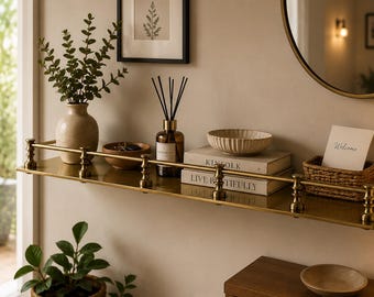 May include: A long, rectangular brass wall shelf featuring a decorative railing along the front edge. The shelf holds a beige vase with greenery, a glass reed diffuser, two stacked books titled Kinfolk and Live Beautifully, and a woven basket.