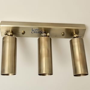 May include: A brass-coloured, three-light fixture with cylindrical shades. The fixture has a rectangular base with the text "Lights Creation Crafts". The shades are adjustable and mounted on the base.