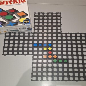 May include: A board game box titled "Qwirkle" with colourful geometric shapes. The game board is made of interlocking grey tiles, with various coloured shapes placed on the board. The box shows the game's name and the shapes used in the game.