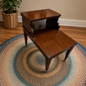 May include: A two-tiered wooden end table with a dark brown finish, featuring a raised upper shelf and a lower main surface. The table is supported by four tapered legs, each finished with a small brass-toned metal caster wheel at the base.