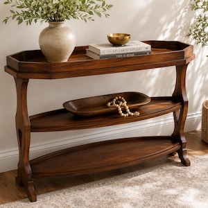 May include: A dark brown wooden console table with three tiers, featuring a raised edge on the top shelf. The table holds a beige ceramic vase, a brass bowl, and two books titled Live Beautifully and This Is Home, plus a wooden bowl with beads.