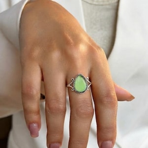 Op de afbeelding: Een zilveren ring met een druppelvormige lichtgroene steen in een gedraaide metalen rand. De ringband heeft een gespleten ontwerp met decoratieve krullen aan beide zijden, wat een gedetailleerd kader vormt rond de centrale edelsteen.