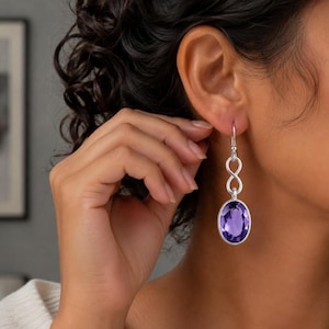 May include: Silver-toned drop earrings featuring a large, oval-cut purple gemstone. The earrings have an infinity symbol design connecting the hook to the gemstone. The gemstone is set in a silver bezel.