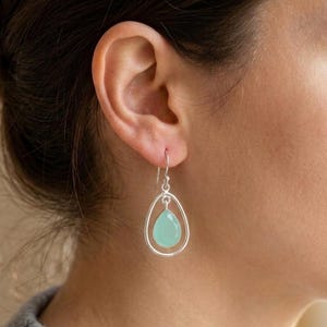 Peut inclure: Une boucle d'oreille pendante en argent avec une pierre bleu clair en forme de goutte, suspendue à l'intérieur d'un cadre ovale ouvert en argent lisse. La boucle d'oreille est fixée à un crochet simple, présentant une finition métallique nette.