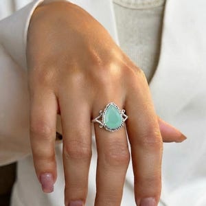 Op de afbeelding: Een zilveren ring met een druppelvormige, lichtgroene steen in een gedraaide metalen rand. De ringband heeft aan beide zijden van de centrale zetting een decoratief, opengewerkt ontwerp, wat zorgt voor een gedetailleerde en gestructureerde uitstraling.
