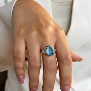 Op de afbeelding: Een zilveren ring met een druppelvormige, doorschijnende lichtblauwe steen in een gedraaide metalen rand. De ringband is aan beide zijden van de centrale zetting voorzien van decoratief metalen krulwerk, wat zorgt voor een gedetailleerd uiterlijk.