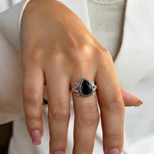 Op de afbeelding: Een zilveren ring met een centrale peervormige zwarte steen in een gedraaide metalen rand. De ringband is aan beide zijden van de zetting voorzien van decoratieve kruldetails, wat zorgt voor een opvallend contrast tussen de donkere steen en de gepolijste zilveren afwerking.