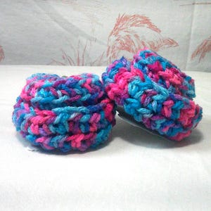 May include: Two round, textured crochet scrubbers featuring a variegated yarn pattern in vibrant shades of bright pink, electric blue, and deep purple. The circular items have a thick, looped construction with a dense, soft, and tactile surface.