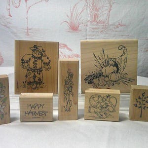 May include: A collection of six light-colored wooden rubber stamps featuring various designs including a scarecrow, a cornstalk, a cornucopia, a tree, a floral vine, and the text Happy Harvest, all arranged in a group on a flat surface.
