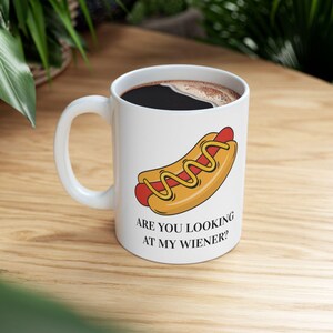 Puede incluir: Taza de café de cerámica blanca con asa, llena de líquido oscuro. La taza presenta un dibujo de un perrito caliente con mostaza y el texto "ARE YOU LOOKING AT MY WIENER?" en la parte delantera. La taza está sobre una superficie de madera.