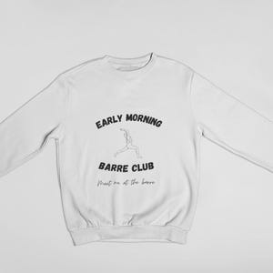 May include: White sweatshirt with long sleeves. The front features the text "EARLY MORNING BARRE CLUB" in black, with a line drawing of a person in a ballet pose. Below the text is the phrase "Meet me at the barre" in a script font.