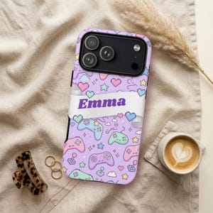 May include: A light purple phone case featuring a repeating pattern of pixelated video game controllers, hearts, stars, and clouds. A white horizontal banner across the center displays the name Emma in bold purple sans-serif lettering.