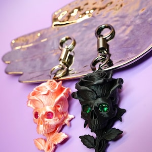 May include: Two keychains featuring rose-shaped skulls. One is peach-colored with pink gem eyes, the other is black with green gem eyes. Both have silver-tone key rings and are displayed on a decorative silver-toned tray against a purple background.