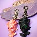 May include: Two keychains featuring rose-shaped skulls. One is peach-colored with pink gem eyes, the other is black with green gem eyes. Both have silver-tone key rings and are displayed on a decorative silver-toned tray against a purple background.