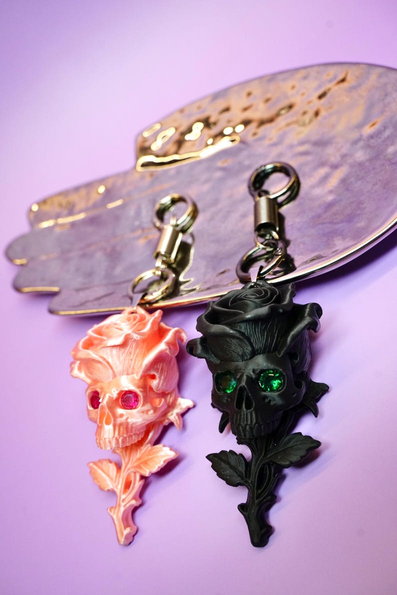 May include: Two keychains featuring rose-shaped skulls. One is peach-colored with pink gem eyes, the other is black with green gem eyes. Both have silver-tone key rings and are displayed on a decorative silver-toned tray against a purple background.