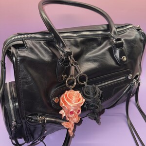 May include: Black leather handbag with silver zippers and hardware. Two keychains hang from the handle: a peach-colored skull rose and a black rose. The bag has a top handle and a shoulder strap.