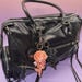 May include: Black leather handbag with silver zippers and hardware. Two keychains hang from the handle: a peach-colored skull rose and a black rose. The bag has a top handle and a shoulder strap.