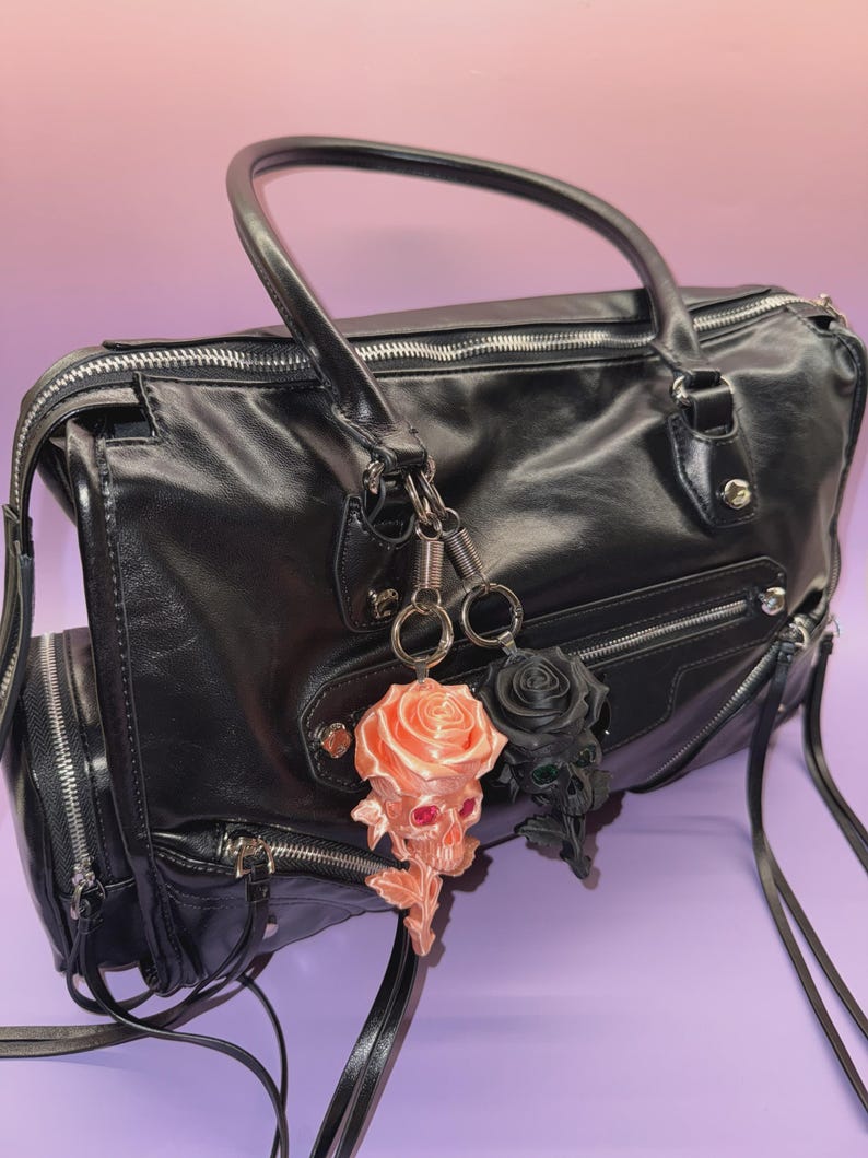 May include: Black leather handbag with silver zippers and hardware. Two keychains hang from the handle: a peach-colored skull rose and a black rose. The bag has a top handle and a shoulder strap.