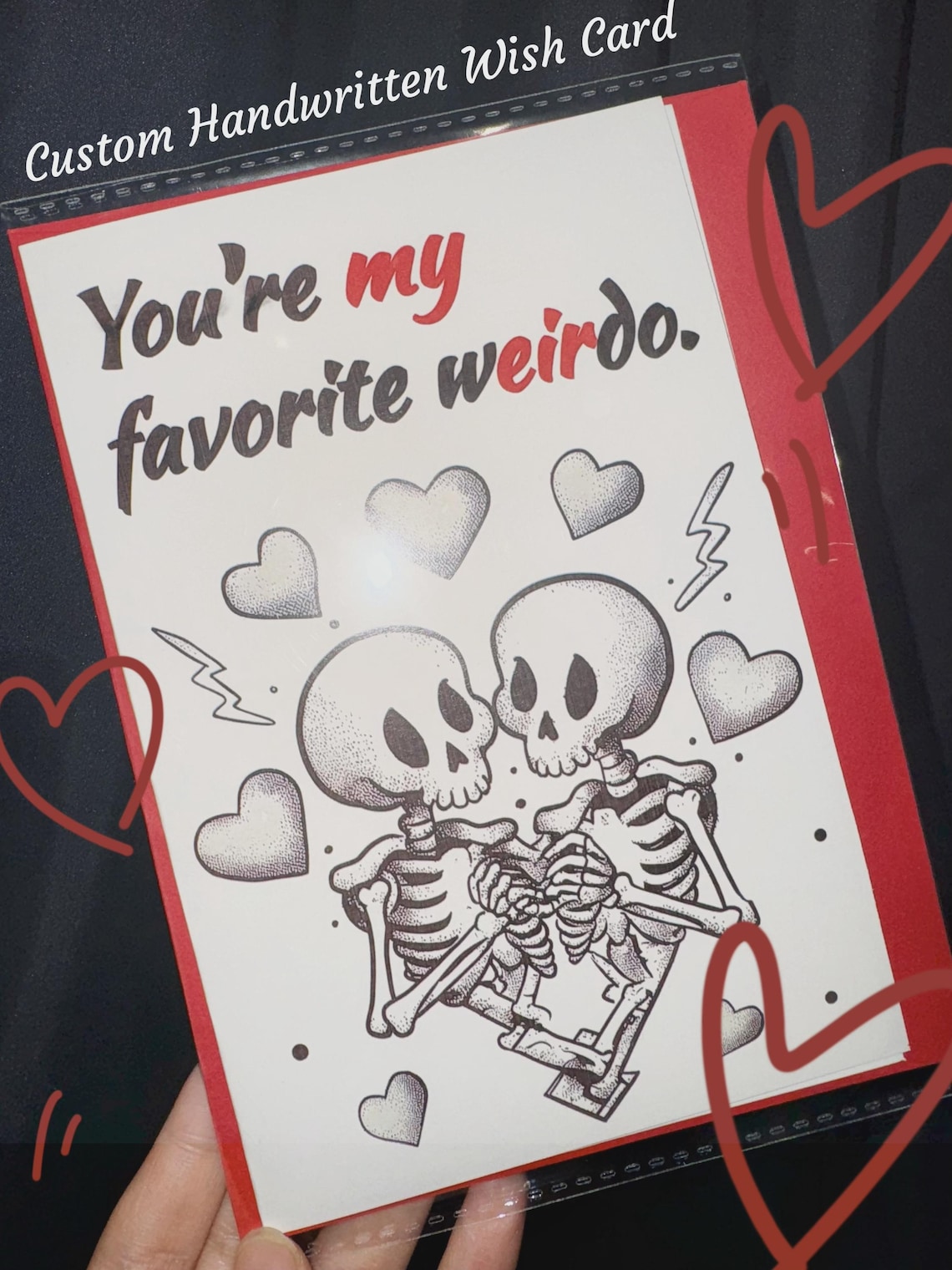 May include: Custom handwritten wish card with the text "You're my favorite weirdo." The card features a black and white illustration of two skeletons embracing, surrounded by hearts and lightning bolts, with a red border.
