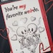 May include: Custom handwritten wish card with the text "You're my favorite weirdo." The card features a black and white illustration of two skeletons embracing, surrounded by hearts and lightning bolts, with a red border.