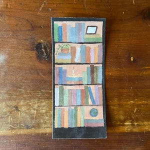 May include: A rectangular bookmark featuring a watercolor painting of a bookshelf. The bookshelf is filled with colorful books, a small potted plant, and a framed picture. The bookmark has a black border and is set against a wooden background.
