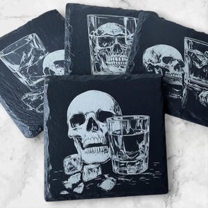 May include: A set of black slate coasters with white etched designs. Each coaster features a skull and a glass of whiskey with ice cubes. The coasters are square and have a natural, textured edge.