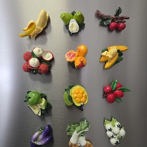 May include: A collection of twelve individual fruit and vegetable magnets arranged in a grid. The set includes a peeled banana, avocado, cherries, lychee, orange, banana bunch, lime, jackfruit, red berries, eggplant, bok choy, and garlic bulbs.