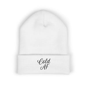 May include: White beanie with the text "Cold AF" in black script across the folded brim. The hat is made of a soft, knitted material and is designed to keep the head warm. The beanie is a casual accessory.