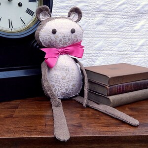 May include: A plush brown and white bear with a pink bow tie. The bear is sitting on a wooden table with a stack of books behind it.