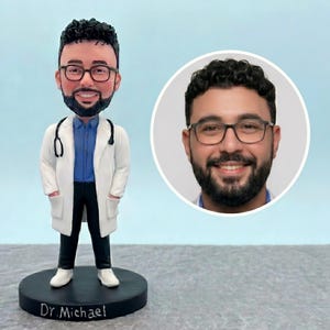 May include: A bobblehead figurine of a doctor wearing a white coat, blue shirt, and black pants, with a stethoscope around the neck. The figurine has glasses and a beard. A photo of the doctor's face is in a white circle. The base reads "Dr. Michael."
