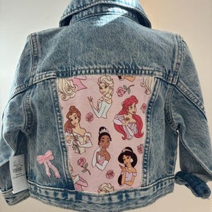 May include: Light blue denim jacket with a pink panel featuring cartoon princesses. The jacket has a classic collar, button closures, and a bow applique. The back panel showcases illustrations of various princesses and roses.