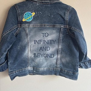 May include: Blue denim jacket with a planet patch and the words "TO INFINITY AND BEYOND" printed on the back. The jacket has a classic collar, button-up front, and long sleeves. White paint splatters add a unique touch to the design.