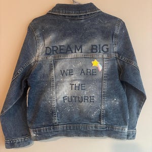 May include: A blue denim jacket featuring a white paint splatter effect on the back. The text "DREAM BIG" is embroidered in dark blue above the text "WE ARE THE FUTURE". A small yellow star patch with a pastel rainbow tail is attached.